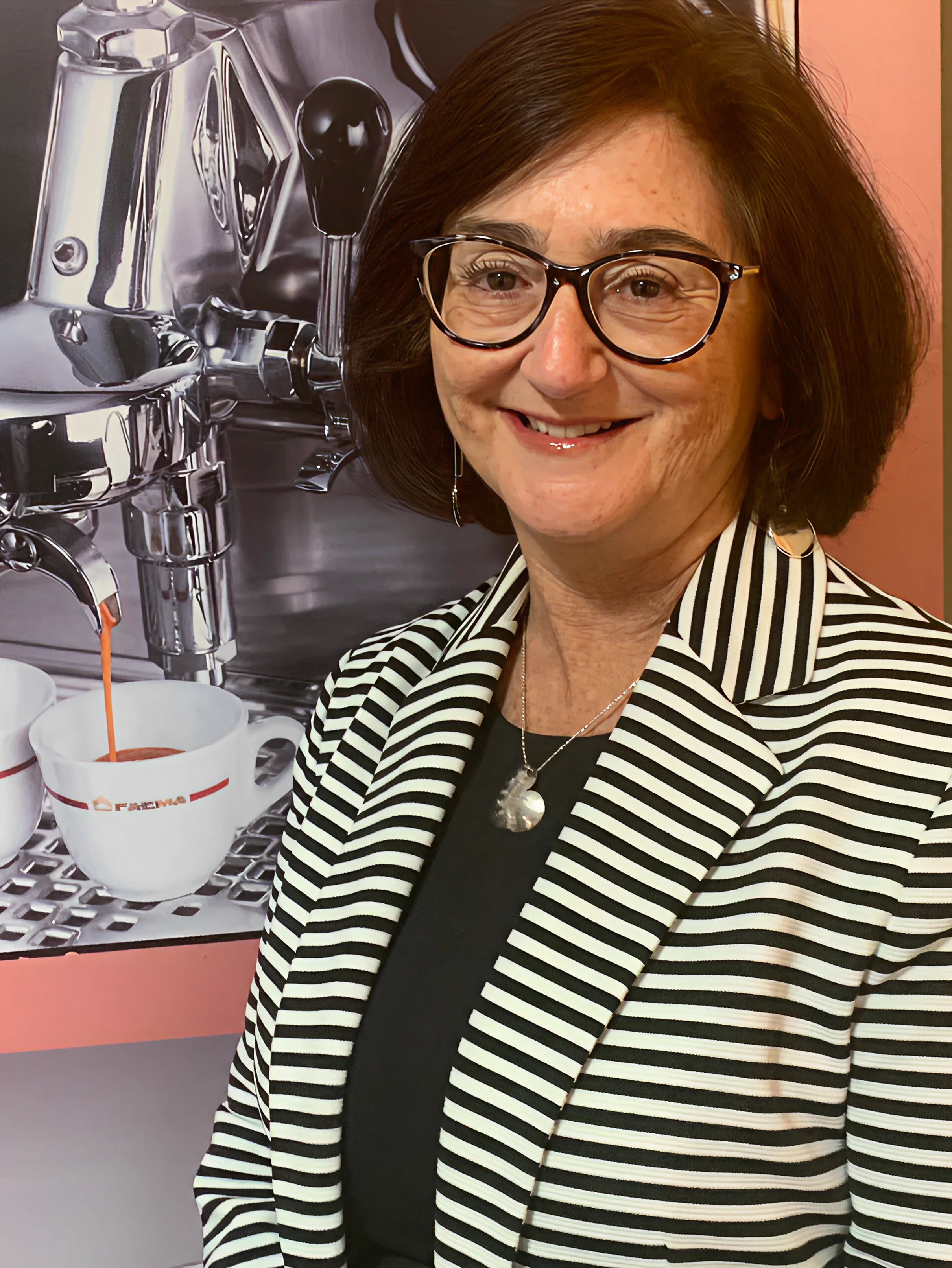 67e57b7677c31_Linda A woman in glasses and striped jacket standing next to coffee machine.